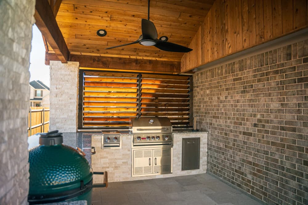 Modern outdoor kitchen featuring a grill, sink, and fan under a wooden ceiling and stone accents.
