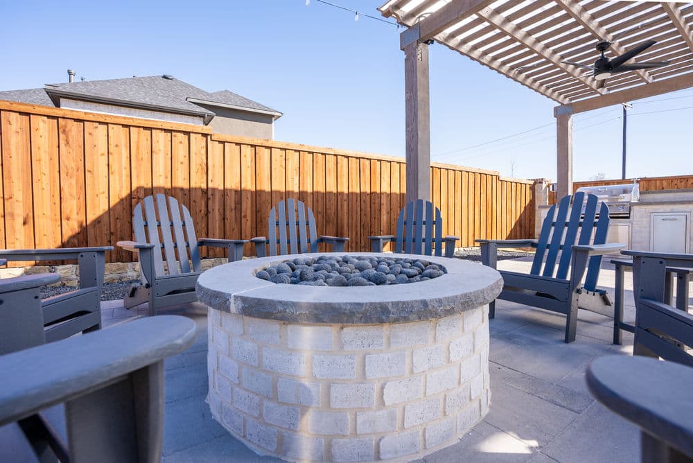 Patio fire pit surrounded by Adirondack chairs in a cozy outdoor setting. Wooden fence in background.